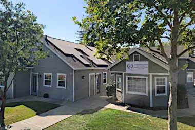 Exterior view of connected residential-style buildings with gray siding, pitched roofs, manicured lawn, and concrete walkway.