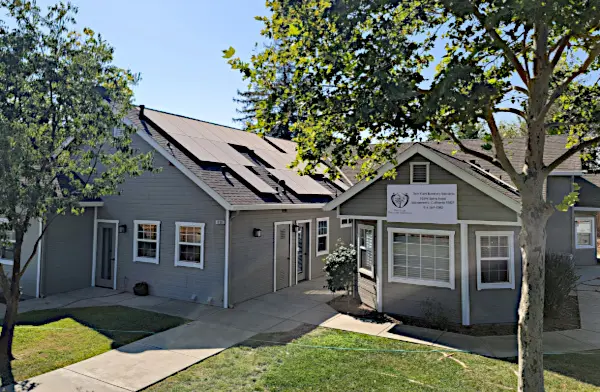 Exterior view of connected residential-style buildings with gray siding, pitched roofs, manicured lawn, and concrete walkway.