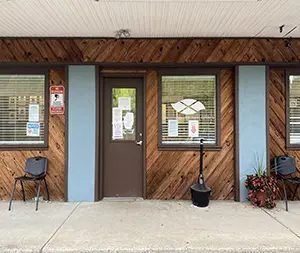 Exterior front facade showing wood-clad entrance, window with logo decal, concrete entry area, and potted flowering plants