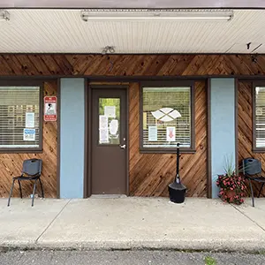 Exterior front facade showing wood-clad entrance, window with logo decal, concrete entry area, and potted flowering plants