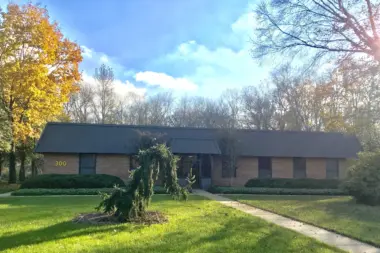Exterior view of one-story brick building with dark metal roof set on green lawn with mature trees and landscaping