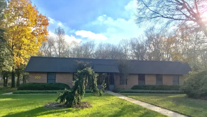 Exterior view of one-story brick building with dark metal roof set on green lawn with mature trees and landscaping