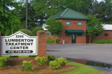 Exterior view of red brick treatment center building with front entrance, green roof, landscaping, and facility monument sign displaying name and phone number.
