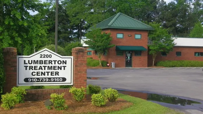 Exterior view of red brick treatment center building with front entrance, green roof, landscaping, and facility monument sign displaying name and phone number.