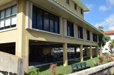 Exterior ground-level view of cream-colored building with large glass windows and doors, landscaping, and seawall.