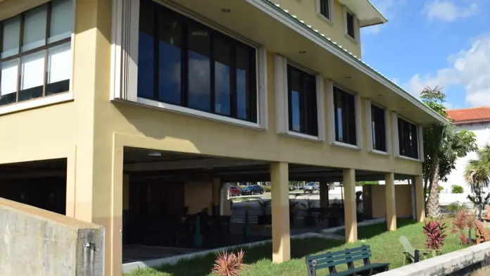 Exterior ground-level view of cream-colored building with large glass windows and doors, landscaping, and seawall.