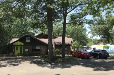 Exterior of single-story brick health clinic building with yellow entrance door and mature tree canopy