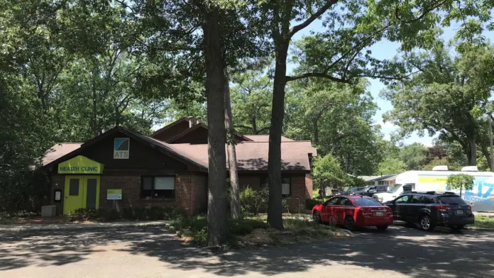 Exterior of brick health clinic building with green accents, parking area with vehicles, mature trees providing shade