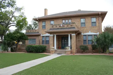 Exterior two-story brick residential building with white columns, covered front porch, landscaped lawn and mature trees