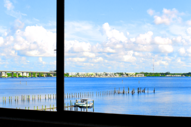 Interior view through window frame showing waterfront landscape, dock pilings, residential buildings, and blue sky.