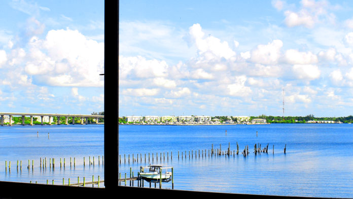 Interior view through window frame showing waterfront landscape, dock pilings, residential buildings, and blue sky.