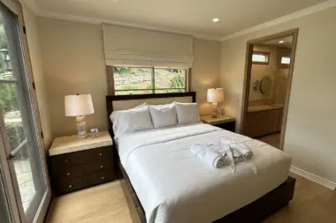 Interior patient bedroom with queen bed, dark wood furniture, ensuite bathroom, and glass door to outdoor terrace with landscaping