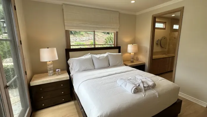 Interior patient bedroom with queen bed, dark wood furniture, ensuite bathroom, and glass door to outdoor terrace with landscaping