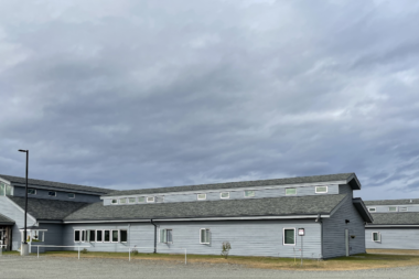 Exterior view of single-story residential treatment facility with light gray siding, multiple windows, covered entry, and parking area.