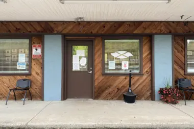 Exterior front entrance of wood-clad treatment facility with brown door, windows, concrete walkway, and potted flowers