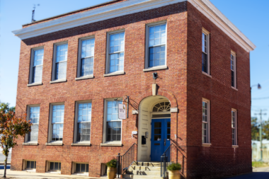 Exterior three-story brick building with blue entrance door, metal railings, multiple windows, and landscaping.