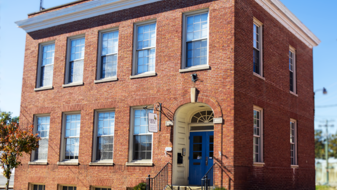 Exterior three-story brick building with blue entrance door, metal railings, multiple windows, and landscaping.