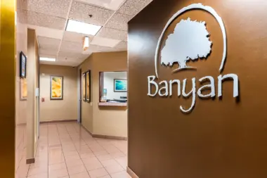 Interior corridor hallway with tan walls, framed artwork, service window, and Banyan Treatment Centers logo on accent wall.
