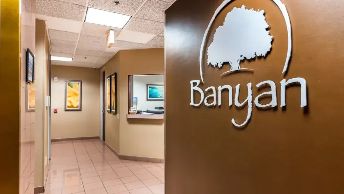 Interior corridor hallway with tan walls, framed artwork, service window, and Banyan Treatment Centers logo on accent wall.