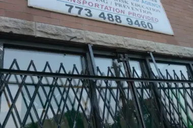 Exterior brick facade showing facility signage with service offerings and phone number, black security gate over windows and entrance.