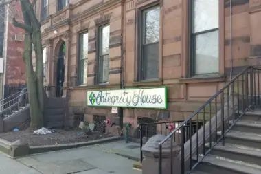 Exterior front entrance of Integrity House brownstone with green neon sign, metal railings, and entry stairs