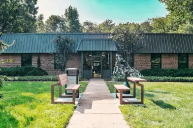 Exterior front entrance showing main doors under covered walkway with landscaping and sitting benches