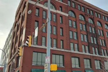 Exterior view of multi-story red brick building with regular windows, green awnings, and street-level entrance in urban setting.