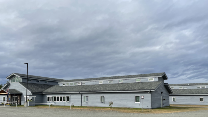 Exterior view of single-story treatment facility with light gray siding, multiple windows, entrance area, and parking lot under cloudy sky
