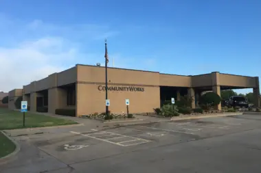 Exterior view of Community Works building with tan brick facade, parking lot, landscaping, and main entrance signage