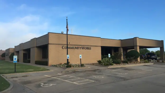 Exterior view of Community Works building with tan brick facade, parking lot, landscaping, and main entrance signage