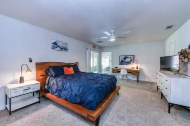 Interior bedroom with wooden bed frame, blue bedding, white furniture, desk area, and glass door to patio