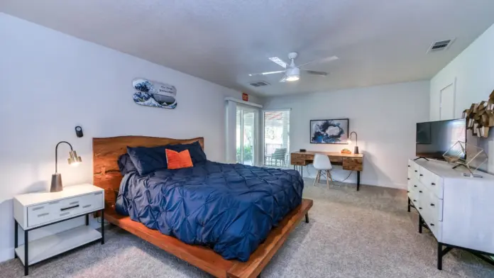 Interior bedroom with wooden bed frame, blue bedding, white furniture, desk area, and glass door to patio