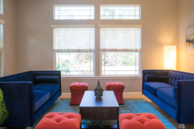 Interior bedroom with upholstered gray headboard, patterned bedding, large windows with shutters, and potted plant