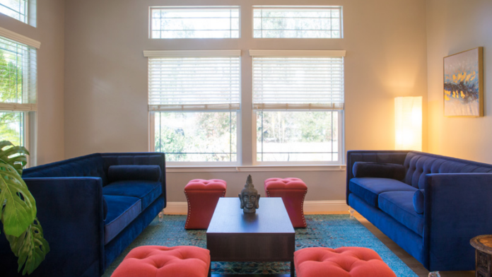 Interior bedroom with upholstered gray headboard, patterned bedding, large windows with shutters, and potted plant
