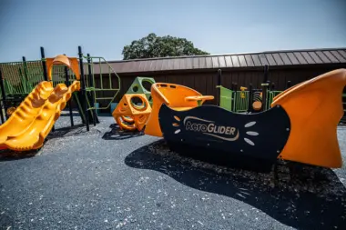 Exterior playground area with orange and yellow climbing structures, slides, and green safety fencing against facility building