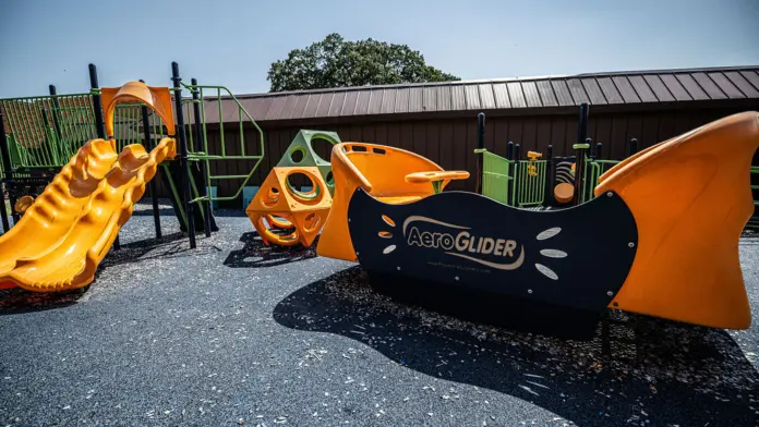 Exterior playground area with orange and yellow climbing structures, slides, and green safety fencing against facility building