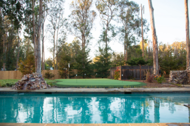 Exterior recreational area with swimming pool, putting green with flagstick, tall eucalyptus trees, and wooden lounge chairs