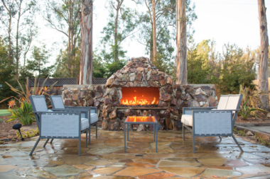 Exterior patio with stone fireplace feature, blue lounge chairs, natural landscaping, and tall trees