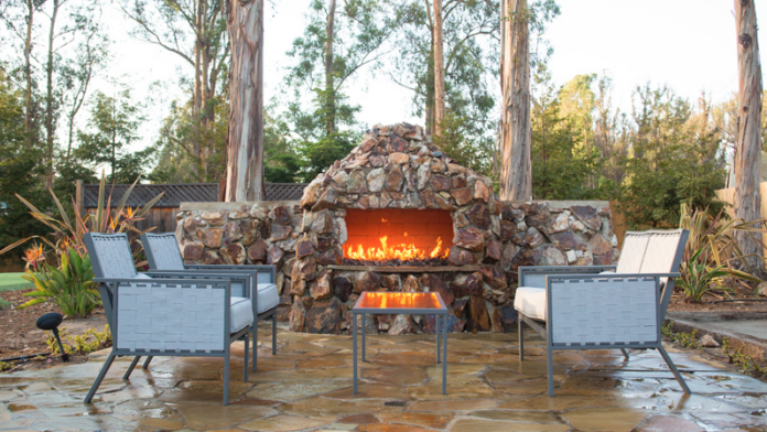 Exterior patio with stone fireplace feature, blue lounge chairs, natural landscaping, and tall trees