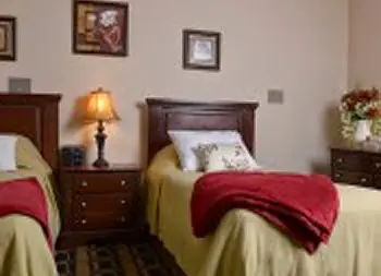 Interior patient bedroom with two beds, wooden furniture, lamps, wall art, and carpet flooring