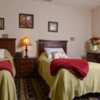 Interior patient bedroom with two beds, wooden furniture, lamps, wall art, and carpet flooring