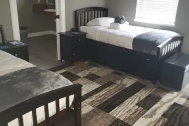 Interior bedroom showing twin bed, nightstand, bathroom entry, window blinds, patterned rug, and dark furniture.