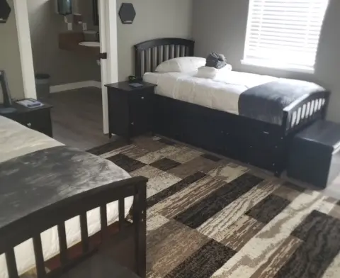 Interior bedroom showing twin bed, nightstand, bathroom entry, window blinds, patterned rug, and dark furniture.