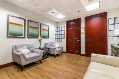 Interior consultation room with cream walls, framed artwork, gray chairs, wooden doors, and wooden flooring.