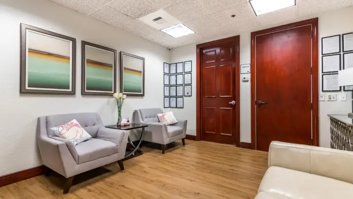 Interior consultation room with cream walls, framed artwork, gray chairs, wooden doors, and wooden flooring.