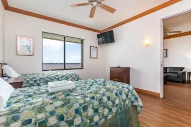 Interior bedroom with twin beds, patterned bedding, wooden ceiling beams, wall-mounted television, and waterfront window views.