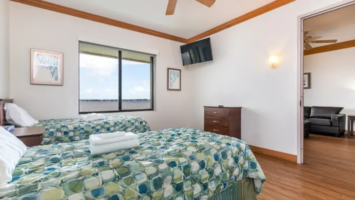 Interior bedroom with twin beds, patterned bedding, wooden ceiling beams, wall-mounted television, and waterfront window views.