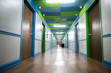 Interior hallway with alternating green and blue framed doors, colored ceiling panels, and polished wood flooring