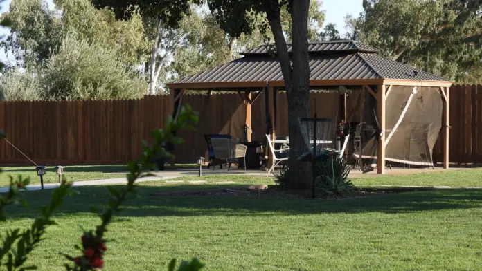 Outdoor patio area featuring wooden pergola shelter, multiple lounge chairs, large tree, wooden fence, and landscaped lawn.
