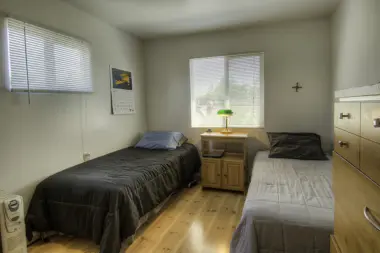 Interior bedroom showing two twin beds, nightstand, windows with blinds, and hardwood flooring